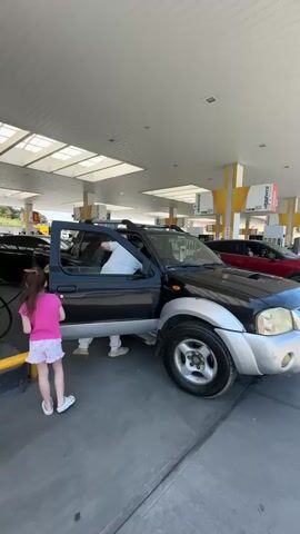 At a gas station, a dark-colored pickup truck is parked at a gas pump. A man in a white shirt is inside the truck on the driver's side, with the door open. A young girl in a pink shirt and patterned shorts stands next to the truck, facing towards the man. Other vehicles and gas pumps are visible in the background. The station has a white roof supported by yellow pillars.