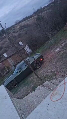 The video shows a view of a dark green Dodge Challenger parked on a grassy area next to a concrete surface. Behind the car is a hillside with some houses. The camera then moves onto the concrete surface, which has various debris scattered on it including tree branches and an empty plastic water bottle. There is also a metal framework in the background. The sky is overcast.