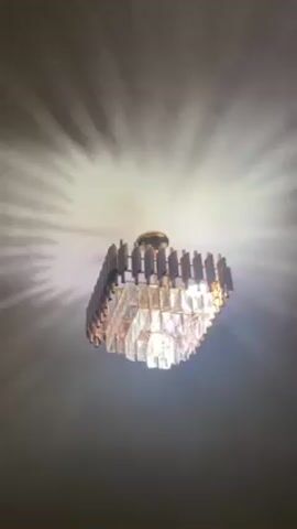 The video shows a chandelier hanging from a ceiling. The chandelier has a square shape and is made of gold-colored metal and clear crystals. The light from the chandelier casts a radial pattern of shadows on the ceiling. The camera is pointed up at the chandelier from below.
