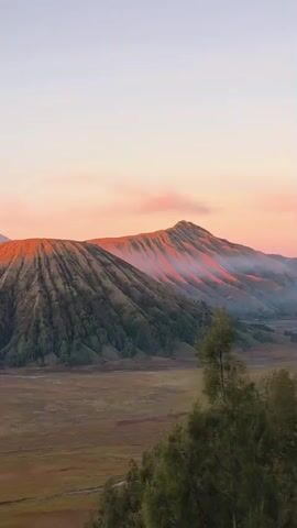 A panoramic drone shot pans across a vast, open landscape dominated by several large volcanoes. The sky is a soft gradient of pale orange and pink, indicating either sunrise or sunset. In the foreground, dry, golden-brown grass covers the ground, with a few sparse trees dotting the terrain. The middle ground features a wide, flat plain with some water features or dry riverbeds. The main focus of the video is the majestic volcanoes. The closest volcano is a conical peak covered in green vegetation, with deep ridges running down its sides. Its summit is bathed in the warm glow of the sky, making it appear orange. To the left and slightly behind this volcano, another larger volcano is visible, with a more rugged and barren appearance. Smoke or steam emanates from its crater and billows into the air, tinged with the colors of the sky. Further in the distance, more volcanic peaks are discernible, their slopes illuminated by the same celestial light. The overall impression is one of natural grandeur and serene beauty.