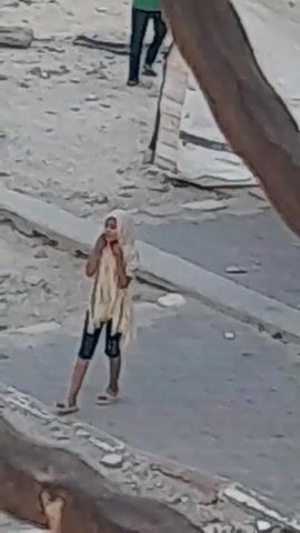 A young person wearing a cream-colored headscarf and a cream-colored garment over a patterned t-shirt and dark shorts walks across a street. The street has white stripes indicating a pedestrian crossing. The environment appears to be dusty and arid, with debris on the ground and what looks like a rudimentary structure in the background.