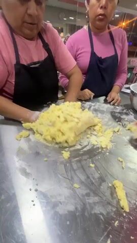 The video shows two women in aprons and hair coverings kneading a large ball of yellow dough on a stainless steel table dusted with flour. The woman on the left is actively working the dough with both hands, while the woman on the right stands behind her, observing. Another person's hand, holding a red spatula, is visible on the right side of the frame, seemingly assisting or preparing to assist with the dough. The environment appears to be a kitchen or food preparation area, with a blurred background suggesting other people and possibly shelves or equipment. The lighting is somewhat dim, with artificial lights visible.