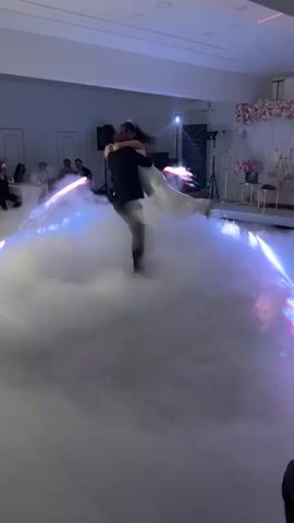 A bride and groom share their first dance on a white tile dance floor in a wedding venue. The room is decorated with a floral display against a white wall, with additional seating to the side. A DJ stand and other music equipment are visible. The couple is surrounded by a low-lying fog effect, lit with blue and white lights. Sparklers are ignited on either side of them, adding to the festive atmosphere. They hold each other as the groom twirls his bride. Some guests are seated at nearby tables. A cameraman stands by the platform at the back of the room.