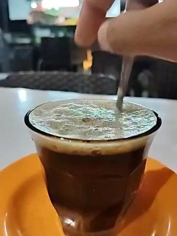 The video shows a cup of dark coffee on an orange saucer, being stirred with a small spoon. The coffee has a frothy surface. The spoon is held in a person's hand. The coffee is in a clear, faceted glass. The background is blurry, but it appears to be a restaurant or cafe setting with tables and chairs visible. The surface the cup is resting on is white.