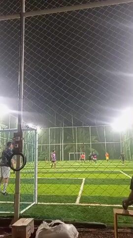 At night, a soccer game is in progress on a field enclosed by a fence and lit by bright overhead lights. The field is artificial turf, with white lines marking the boundaries. Several players wearing brightly colored jerseys can be seen actively playing. One team is wearing orange jerseys and the other is wearing purple and blue. A person stands outside the fence, watching the game. Another person is sitting on a stool outside the fence.
