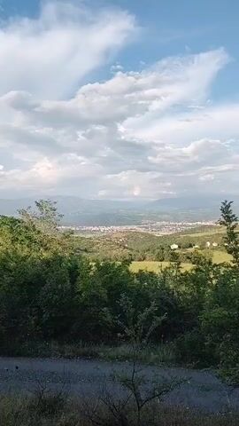 The video captures a panoramic view of a town nestled in a valley, surrounded by mountains and lush green hills, with a bright sky and scattered clouds overhead. The camera pans from left to right, initially showing a wide landscape with the town in the mid-ground. It then zooms in on a prominent, large white cross situated on a distant, forested mountain, overlooking the town. The town features numerous multi-story buildings, typical of urban development. The foreground consists of trees and shrubs, suggesting the video was shot from an elevated, possibly rural or semi-rural, vantage point. The scene appears peaceful and well-lit, indicating daytime conditions, likely late morning or afternoon. No visible text is present in the scene. The location context provided is Selishte, Bulgaria.