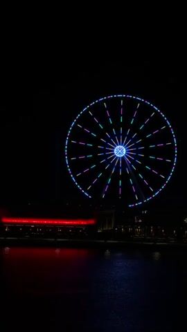 A large Ferris wheel is illuminated with colorful lights against a black night sky. Below the Ferris wheel, a waterfront area is visible with a red-lit building in the background and dark water in the foreground reflecting the lights. The Ferris wheel appears to be slowly rotating, as evidenced by the slight shift in the positions of its lights over the short duration of the video. There are no people or animals visible in the frame.