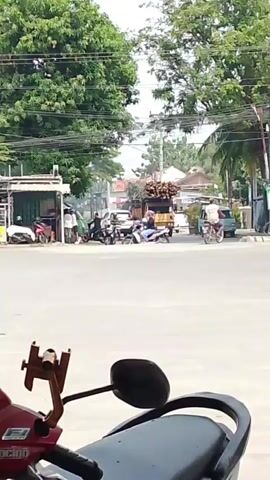The video shows a street intersection with several vehicles and people. In the foreground, the seat and rear-view mirror of a red motorcycle are visible. In the background, a yellow truck loaded with wood is driving through the intersection. To the left of the truck, a black car is stopped. To the right, a blue van is moving. Several motorcycles and people are also visible around the intersection, with some standing and others riding motorcycles. The scene appears to be in a tropical or subtropical environment, with lush green trees lining the street and power lines overhead. The sky is overcast, suggesting it may be a cloudy day.