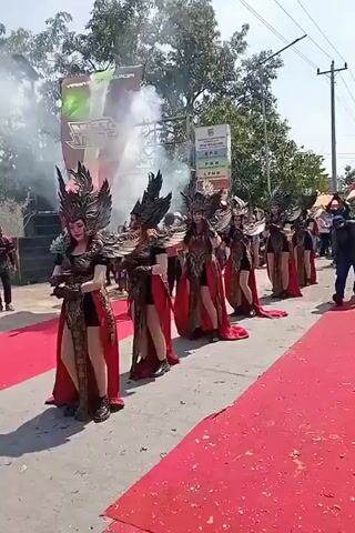 A group of seven women, dressed in elaborate, winged, dragon-like costumes with gold and red accents, walk in a line down a red carpet. They are carrying flaming torches that emit white smoke. Behind them, other people are visible, along with some structures and trees. The scene appears to be outdoors, possibly at a festival or parade, with a clear sky.