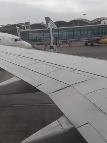 The video shows a view from the wing of an airplane, with another plane parked on the tarmac nearby. The parked plane is an Air Algérie Boeing 737, painted white with red accents. The engine is visible, and the registration number on the tail is 7T-VKJ. In the background, there is a covered walkway connecting the terminal to the aircraft. The weather appears overcast, and the tarmac is a gray concrete surface. The video appears to have been taken from inside the airplane, at a gate.