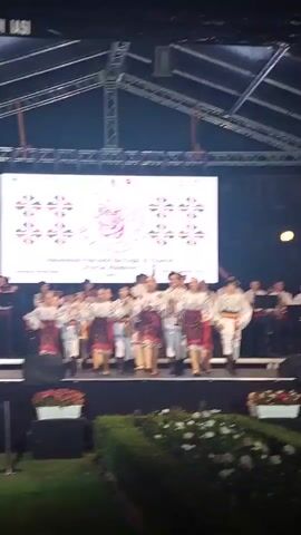 A group of children and teenagers dressed in traditional Romanian folk costumes are performing on a stage in front of a large screen. The screen displays the words "CATALINA" and "Ansamblul Folcloric de Copii si Tineret Florile Moldovei Iasi" above a decorative pattern. Some of the performers are holding musical instruments. There are also adults playing instruments on stage. The stage is set up outdoors under a tent-like structure. In the foreground, there are bushes and flowers.