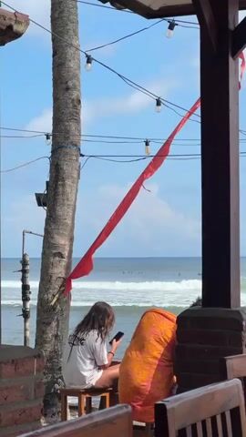 Two women sit on stools on the porch of a beachfront building, looking at the ocean and presumably interacting with each other. One woman has dark hair, a white T-shirt, and is looking at a smartphone. The other woman has dark hair and is wrapped in an orange and yellow patterned fabric. A tall palm tree stands to the left of the women, decorated with hanging lights and an old red flag. Behind them is the ocean, with multiple surfers in the water. A man in a gray long-sleeved shirt and blue trunks walks past them with a yellow surfboard under his arm. The porch has wooden beams and a brick foundation.