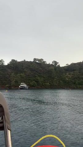 The video shows a view from a boat on the water, looking towards a hilly, tree-covered coastline. Several yachts are visible anchored in the water near the shore. The sky is overcast and the water is choppy.