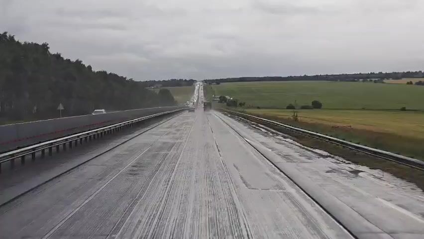 The video shows a drone's eye view of a wet highway. The highway is paved with gray asphalt and has white lane markings. There is a concrete barrier on the left side of the road, and a metal guardrail on the right. The road is surrounded by green fields, with some trees and a few buildings in the distance. The sky is cloudy and gray, suggesting it might be raining or has recently rained. There are a few vehicles on the road, including cars and trucks. The drone is moving forward along the highway.