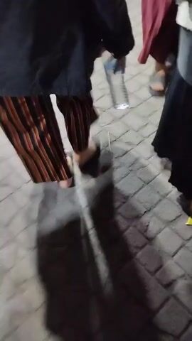 The video shows a crowded outdoor scene at night, possibly a market or gathering. Several people, including women and children, are visible, some sitting on the ground, others standing or walking. They appear to be dressed in casual clothing, with some women wearing headscarves. There are various objects scattered around, including bags, containers, and what looks like food stalls or vendors. The lighting is dim, and the overall atmosphere seems busy and somewhat chaotic. The ground is paved with interlocking stones. The camera movement is shaky, indicating that the video was likely recorded by someone moving through the crowd.
