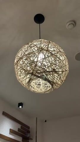 The video pans across a room with white textured walls and a white ceiling. Several decorative items are visible. A large, spherical pendant light made of woven material hangs from the ceiling, illuminated from within. Two smaller, square black ceiling lights are also visible. A wall features a decorative arrangement of horizontal wooden planks against a white textured surface, with what appears to be a small shelf or ledge integrated into the design. Above this, two framed pictures hang on the wall; one shows sliced oranges and a circular dessert, and the other depicts a rolled cake. A twisted, dried tree branch is positioned in the foreground, with a small plant attached to it and another pendant light hanging from it, also made of woven material. The overall aesthetic suggests a modern, possibly cafe or restaurant setting with a natural, artistic theme.