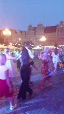 The video shows a group of people dancing in a square at dusk. The ground is paved with cobblestones, and colorful lights are projected onto the surface. In the background, there are buildings with illuminated windows and ornate streetlights. People are holding hands and dancing in a circle. Some individuals are wearing casual clothing like t-shirts and shorts, while others are dressed in more formal attire. The atmosphere appears festive and lively, with a mix of adults and possibly children participating in the dancing. The sky is a deep blue, indicating the transition from day to night.