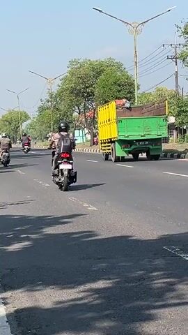 A motorcycle with a rider wearing a black helmet and backpack is traveling on a paved road. The rider is looking forward. A lime green dump truck with a red and orange cargo bed is also on the road. The truck has writing on the side in what appears to be Indonesian. In the background, a yellow dump truck with a green cargo bed is moving away from the camera. Several trees are visible along the side of the road, as well as a lamppost. The sky is clear and blue. The scene appears to be outdoors, during the daytime.