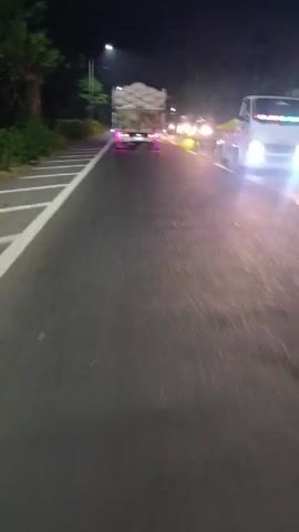 The video presents a nighttime scene from the perspective of a driver on a paved road. The road is marked with a solid white line, indicating lane divisions. Streetlights illuminate the environment. Ahead, a truck with a load covered in a material resembling grey fabric is visible. The truck also features pink underglow lighting. The camera appears to be shaking slightly, suggesting movement of the vehicle. The right side of the road is bordered by vegetation. Several vehicles with illuminated headlights are also observed further down the road.