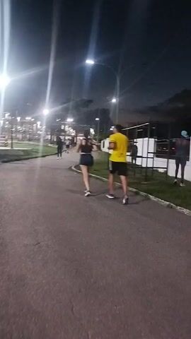 At night, a man and a woman are running along a paved path in a park. The woman is wearing dark shorts and a tank top, while the man is wearing a yellow shirt and dark shorts. Several streetlights illuminate the scene. There are other people walking or standing on the park grounds.