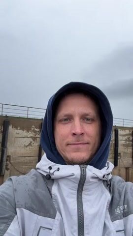 A man with blue eyes and a light beard, wearing a blue hoodie under a grey and white rain jacket, takes a selfie video. He looks directly at the camera with a calm, slight smile. The jacket has "FITE 1997" visible on the lower right.

The background changes as the video progresses, suggesting movement. Initially, a concrete wall or dock structure with black fenders and a body of water are visible under an overcast sky. Later, a distant bridge spanning the water becomes visible, along with parts of the vessel's deck, including a white truck. Finally, red trucks and other vehicles are seen on the ferry deck behind him. The scene suggests he is on a ferry crossing a body of water in Nizhneleninskoe, Russia, during an overcast day.