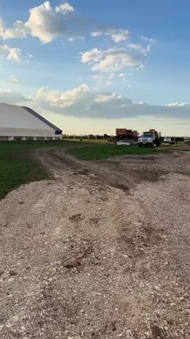 A brown horse trots across a grassy area in front of a large white and blue tent. In the background, there are several vehicles, including a pickup truck and a trailer. The sky is blue with scattered white clouds, and the sun is low, suggesting it is either early morning or late afternoon. The ground in the foreground is a dirt and gravel path.