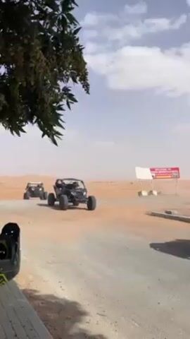 The video shows two black off-road buggies driving up to a paved road in a desert environment. The camera is positioned to the side of the road, under the shade of a tree, and shows the buggies coming toward the viewer. The sky is partially cloudy and a bit hazy, possibly due to the desert dust. A sign with writing on it is visible in the background. The vehicles appear to be stopping or slowing down near the road. There are no visible people outside of the buggies.