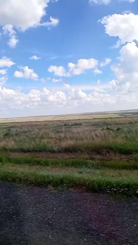The video shows a wide, open landscape under a bright blue sky filled with white, puffy clouds. The foreground is a dark, gravelly surface, possibly a road or path. Beyond the gravel, there is a strip of vibrant green grass, which transitions into a vast expanse of dry, golden-brown grasses and low-lying vegetation. The horizon is flat and stretches across the frame, meeting the sky in a clear line. There is a faint indication of a dirt track or path running horizontally through the grassy area. The overall impression is one of a rural, possibly arid, environment on a sunny day. No people or animals are visible.