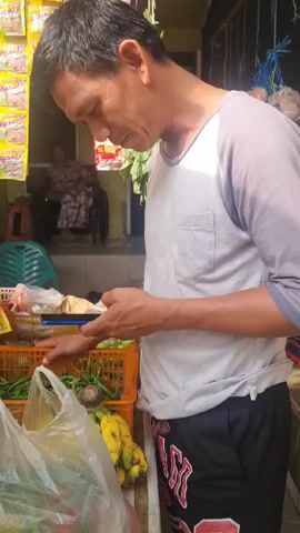 Here is a concise description of the video:

A man stands outdoors at a vegetable stand or stall, looking at his smartphone while holding the handles of a plastic bag containing vegetables. A variety of fresh greens are piled on the stall's surface, including what appears to be bok choy. Behind the counter, there are additional vegetables in baskets and bananas are visible on the counter. A person in a wheelchair is visible in the background near the entrance to a building. The stall is covered in snack items.