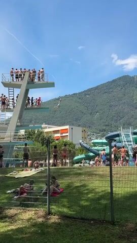 The video shows a busy outdoor swimming pool on a sunny day. In the foreground, people are relaxing on the grass behind a fence. The middle ground features a large diving platform with many people standing on it, some on the higher levels and some on the lower. One person is mid-air, having jumped from the platform. To the right, there are water slides and more people in and around the pool area. In the background, buildings and a large, tree-covered mountain under a clear blue sky can be seen. The overall scene depicts a lively summer day at a recreational facility.