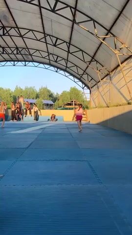 This media file shows an outdoor gathering of people under a large, arched, open-air structure with a metal frame and a translucent roof. The ground is covered with blue, interlocking mats, suggesting a sports or recreational area. Several individuals, including children and adults, are scattered across the mats and on a raised platform in the background.

In the foreground, a young girl in an orange top and shorts is walking towards the left. To her right, another child in a pink outfit is running. Further back, a group of people are standing and sitting on a tiered platform. Some are dressed casually, while others wear more distinctive attire, such as a long, flowing red skirt. A person in the middle distance is sitting on the blue mats in a cross-legged pose.

The scene appears to be taking place during daylight, likely late afternoon or early evening, as evidenced by the warm, golden light casting long shadows. The background reveals lush green trees and some buildings, including structures with blue and wooden roofs. A glimpse of a larger, ornate building with a green turret is visible in the far distance, hinting at a possibly rural or park-like setting. The overall atmosphere seems relaxed and social, with people engaged in various informal activities. The location context provided is Krasnoturansk, Russia.