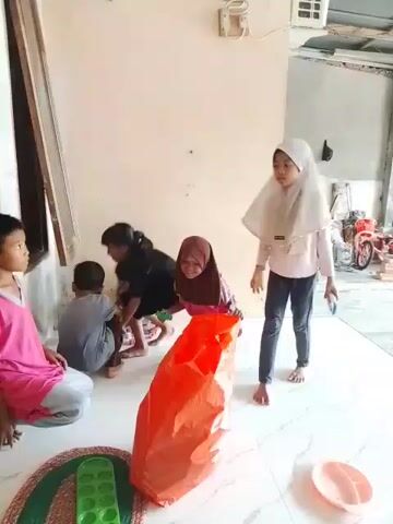 Six children are in an indoor area. A boy wearing a pink shirt sits on the floor in the foreground left, facing away from the camera. To his right, a girl wearing a black shirt and a hijab is also on the floor, facing forward. Another child in a pink shirt is crouched behind her. Two children in the background are on the floor, one in a grey shirt and the other in a pink shirt. To the right, a girl wearing a white hijab and jeans stands and faces away from the camera. An orange bag is on the floor between the children. The environment appears to be a covered outdoor area or a garage with a concrete floor and an unfinished wall.