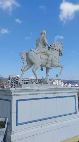 The video pans across an outdoor plaza in front of a large, luxurious multi-story building. The architecture features a pale yellow facade accented with white columns and balustrades. Statues of men and horses decorate the plaza, which also includes pools of water, staircases, and potted plants. The sky is blue with scattered clouds. The video gives a sense of opulence and grandeur.