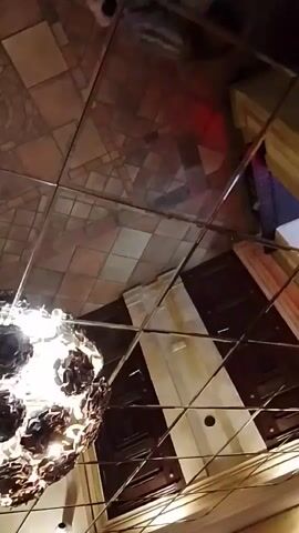 The video shows a blurry, low-angle view of a chandelier with many lights, set against a background of dark, checkered walls and a tiled ceiling reflecting the scene.  The chandelier is the brightest element in the shot. The camera seems to be moving.
