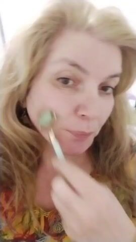 A woman with blonde hair is holding a jade roller to her face and rolling it. She is wearing a colorful shirt. The video is short, showing her face from the shoulders up.