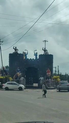 The video shows a large, dark stone gateway with several statues on top. The statues depict figures in historical attire, some holding trumpets and one appearing to be playing a drum. Below the gateway, there are two elephant statues, each with a rider. One elephant is positioned to the left of the gateway and the other to the right. In front of the gateway, there are metal railings and a few human statues. A white car is parked to the left of the gateway, and a white car is driving past on the right side of the road. A person is walking across the street in the foreground. The sky is overcast.