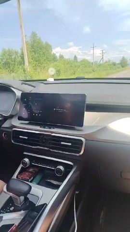 The video shows the interior of a car. The camera is positioned to show the dashboard and the view through the windshield. The dashboard has a large touchscreen display in the center, an instrument cluster behind the steering wheel, air vents, and a gear shifter. A person's hand is visible on the steering wheel, and their legs are visible in the lower part of the frame, suggesting they are the driver. The car is on a paved road, with green trees and vegetation on either side. In the distance, other cars are visible on the road. The sky is partly cloudy. The touchscreen display shows a media player interface.