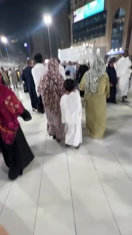 A nighttime outdoor scene shows a large crowd of people walking across a paved area. The people are wearing a variety of clothing, including traditional religious attire like thobes and hijabs, as well as more casual modern clothing. Many are wearing white garments, suggesting a religious pilgrimage. In the background, tall buildings with illuminated windows are visible, and streetlights illuminate the area. The atmosphere appears to be one of movement and congregation.
