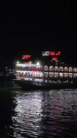 At night, a multi-level boat, lit up with numerous lights, is on the water. The reflection of the lights shimmers on the water. The boat is labeled “SQ Cables” and has other signage in what appears to be the Bengali language. People are visible on the boat.