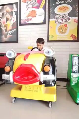 A young child sits in a yellow kiddie ride shaped like a cartoon car with large red lips and googly eyes. The child is looking down at the steering wheel. The kiddie ride is in front of a brick wall with three framed posters advertising food. To the right of the kiddie ride, a green kiddie ride can be seen. The environment appears to be an indoor entertainment area or restaurant.
