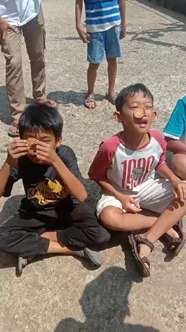 Two young boys sit cross-legged on a concrete surface, each with a cookie placed on their forehead. They are participating in a game, attempting to move the cookie from their forehead to their mouth without using their hands. A group of people, mostly children, stand around them, observing. The boy on the left wears a black t-shirt, the one on the right a red and white baseball shirt. The background is a mix of sunlight and shadows on the concrete surface.