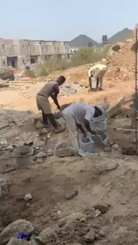 The video shows three people constructing a wall. Two of the people are squatting down working with blocks, while the third person is on a small hill behind them with a shovel. The area is dusty, with scattered rocks, cement blocks, and tools. In the background are rows of unfinished buildings and hills.