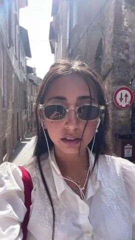 A woman with long, dark hair and sunglasses is walking down a cobblestone street. She is wearing a white shirt and has earbuds in. The street is lined with old buildings, some with arched doorways. There is a traffic sign on the right side of the street. The sun is shining.