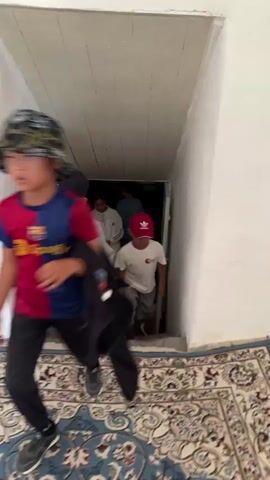 A group of four young people descend a narrow, unfinished concrete staircase. The person filming is moving backwards, following the others down. In the foreground, a person wearing a white hoodie with "USA" on the leg and a blue baseball cap walks away from the camera, their back turned. To their right and slightly behind, a young girl in a white tracksuit with black stripes on the sleeves and grey pants walks down the stairs, her face is visible and she is looking down. She is wearing a red baseball cap. Further down the stairs, two more individuals are visible in the dimly lit area at the bottom of the stairs. One is wearing a blue t-shirt with a palm tree design and patterned shorts. The other, to their left, is wearing a dark t-shirt and green shorts and appears to be holding a phone. The walls of the staircase are white and unfinished. The ceiling above is made of wooden planks. A patterned rug is visible in the very bottom right corner of the frame.