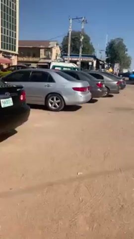 The video shows a row of parked cars on a dusty, unpaved lot under a clear blue sky. The cars are various shades of silver and gray. An ambulance and other vehicles are parked further down the line of cars. Buildings are visible in the background. The camera slowly pans to the right along the row of cars.