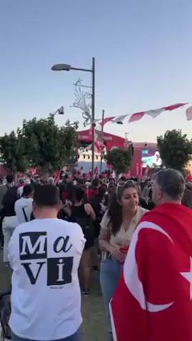 Here is a concise description of the scene:

The video shows a large crowd gathered in a public space, likely a city square, with a stage visible in the background. The crowd appears to be attending an event or gathering. Many individuals are wearing or carrying Turkish flags, suggesting a nationalistic or patriotic theme. The overall atmosphere seems lively, though the specific nature of the event is unclear from the video alone.