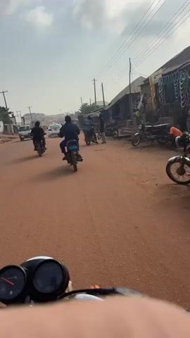 The video shows a street scene from the perspective of someone riding a motorcycle. The road is unpaved and reddish-brown. Several motorcycles, a white car, and two yellow auto-rickshaws are visible. People are riding the motorcycles and walking near the roadside. Buildings line the right side of the street. The sky is cloudy.