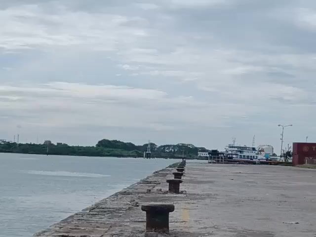 The video shows a concrete pier on a cloudy day. The pier has a series of dark mooring bollards along its edge. To the left, water extends to a distant shoreline with trees and some structures. To the right, a ship is docked along the pier. There's also a street light and a shipping container nearby. The sky is overcast with a mix of light and darker gray clouds.