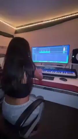 A young woman with long, dark hair is seated in front of a large computer monitor and a keyboard, seemingly in a music production environment. She is wearing a black crop top and grey sweatpants. The monitor displays music editing software with sound waves visible, and the woman appears to be interacting with the keyboard. The room is dimly lit with a string of small lights along the ceiling, creating a warm ambiance. Other objects visible include a computer mouse, studio speakers, and a padded chair. The overall impression is that she is working on a music project.