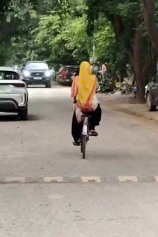 A person on a bicycle, wearing a yellow headscarf and dark pants, cycles down a paved road. There are cars on either side of the road and trees line the background. The person appears to be moving away from the camera.